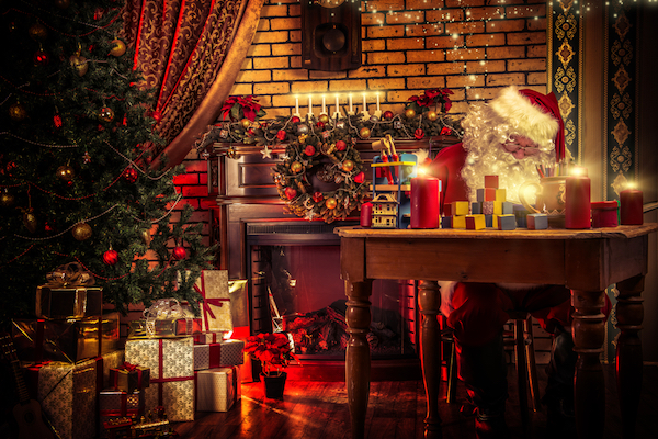 Santa sitting at table with head down. Table covered in candles and toys. Fireplace behind Santa, Christmas tree to his left. Presents beneath tree.